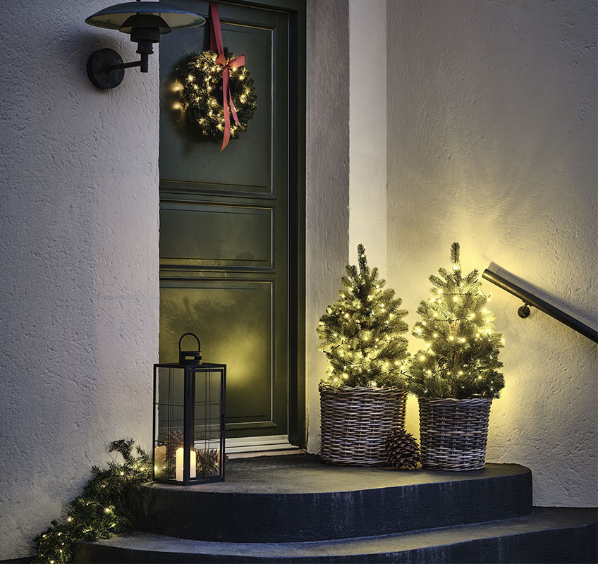 Két világító fa egy piros masnis karácsonyi koszorúval feldíszített bejárati ajtó előtt