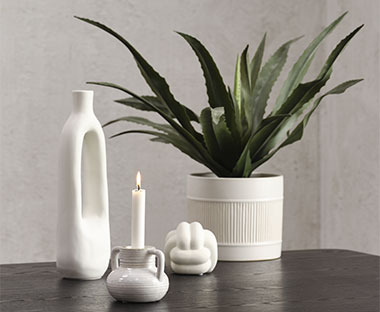 Fehér váza, fehér gyertya gyertyatartóban, fehér kisméretű dekorszobor és műnövény fehér kaspóban egy sötét színű asztalon