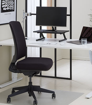 Fekete irodaszék fehér íróasztallal amin fekete monitor állványra helyezett monitor van