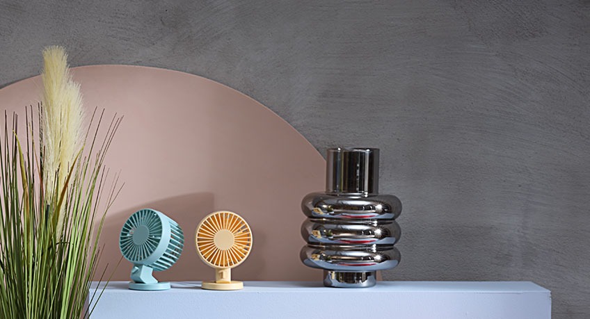 Two mini fans and metal vase beside an artificial plant 
