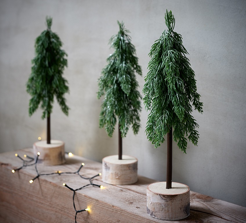 Három műfenyő egy fa talapzaton: modern karácsonyi dekoráció